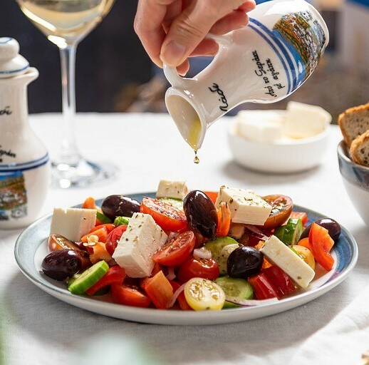 bottle filled with olive oil being poured over a mediterranean salad full of tomatoes, cucumbers, cheese