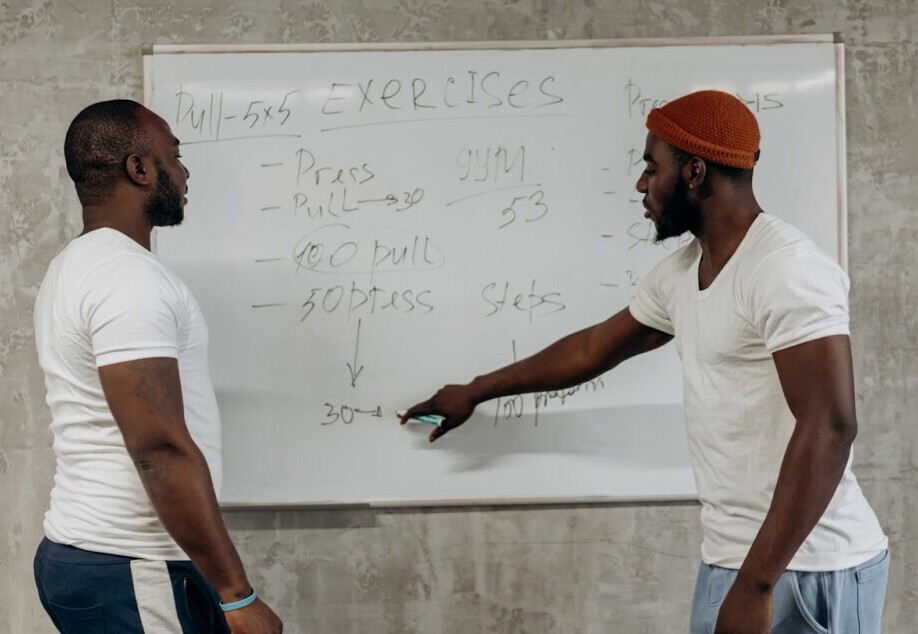 two men in front of a whiteboard with an exercise plan written. one man points to something on the whiteboard for the other man